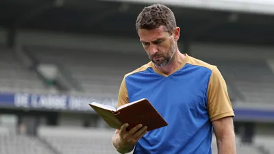 A 35-year-old, 5'10" male coach with a rugged build, stands in a low, blurred background of Stade de l'Abbé Deschamps'...