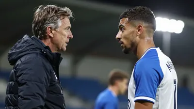 A tight shot of an Auxerre player in a white jersey with blue sleeves, mid-conversation with coach Will Still on the...