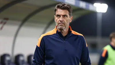 A determined, mid-30s Auxerre coach with a rugged build, wearing a plain blue and amber training top, stands pitchside at...