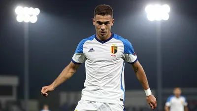 A 21-year-old Belgian midfielder in a white and blue kit, with a clean-cut face and a determined expression, playing a...