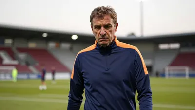 A 50-year-old, 5'10" male coach, dressed in a plain blue and amber polyester training top, his face set in a mixture of...
