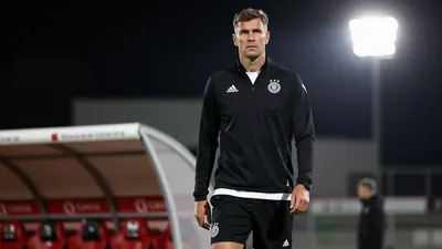 A 28-year-old, 1.80m tall, German coach, wearing a black RC Lens tracksuit, with a white shirt and black shorts, standing...