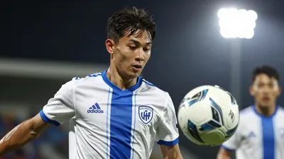 A 22-year-old midfielder in a white and blue jersey, with a blue and white striped shirt underneath, playing a ball with...