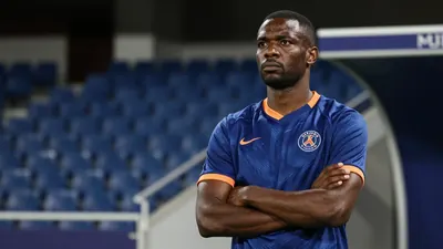 A 35-year-old, 5'10" tall, athletic-build, African-descent, assistant coach, with a stern expression, stands in the...