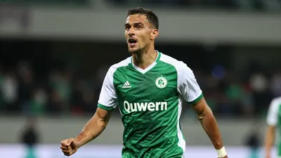 Photorealistic image of an Auxerre player in a green and white jersey, celebrating a late equalizer in a friendly match...